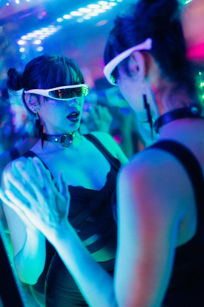 Woman in futuristic sunglasses and outfit, posing with mirror reflection in neon-lit nightclub.